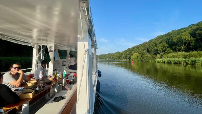 People dining on a boat cruising through a river with trees on the shore.