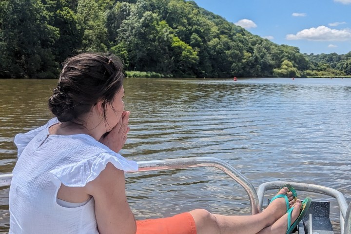 Person sitting on a boat, wearing a white shirt and orange skirt, looking at a river with trees.