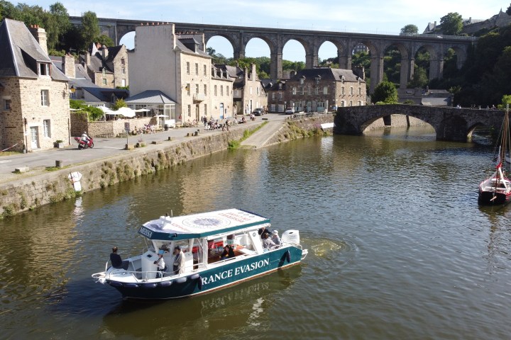 Boat on river with stone bridge and viaduct in background, village buildings on riverbank.