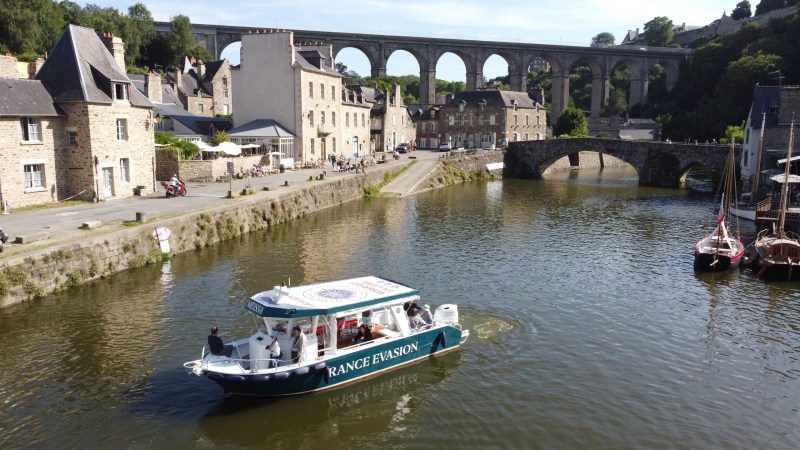 Boat on river with stone bridge and viaduct in background, village buildings on riverbank.