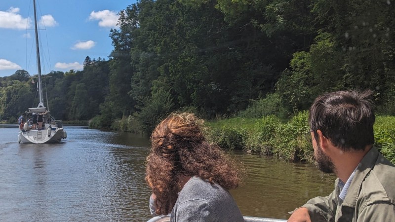 Two people on a boat observing another boat on a calm river.