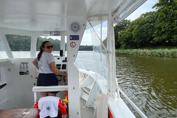 Person steering a boat on a river with lush green banks.