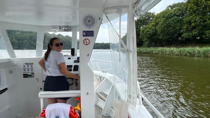 Person steering a boat on a river with lush green banks.