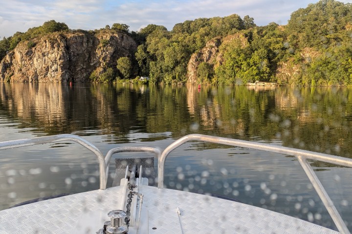 Boat deck on calm water with rocky, tree-lined shore in the background.
