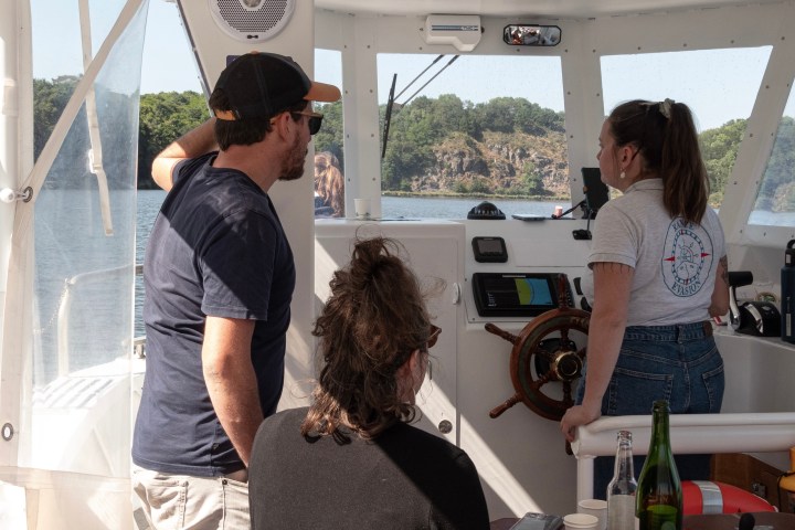 Three people on a boat, one steering, with water and trees in the background.